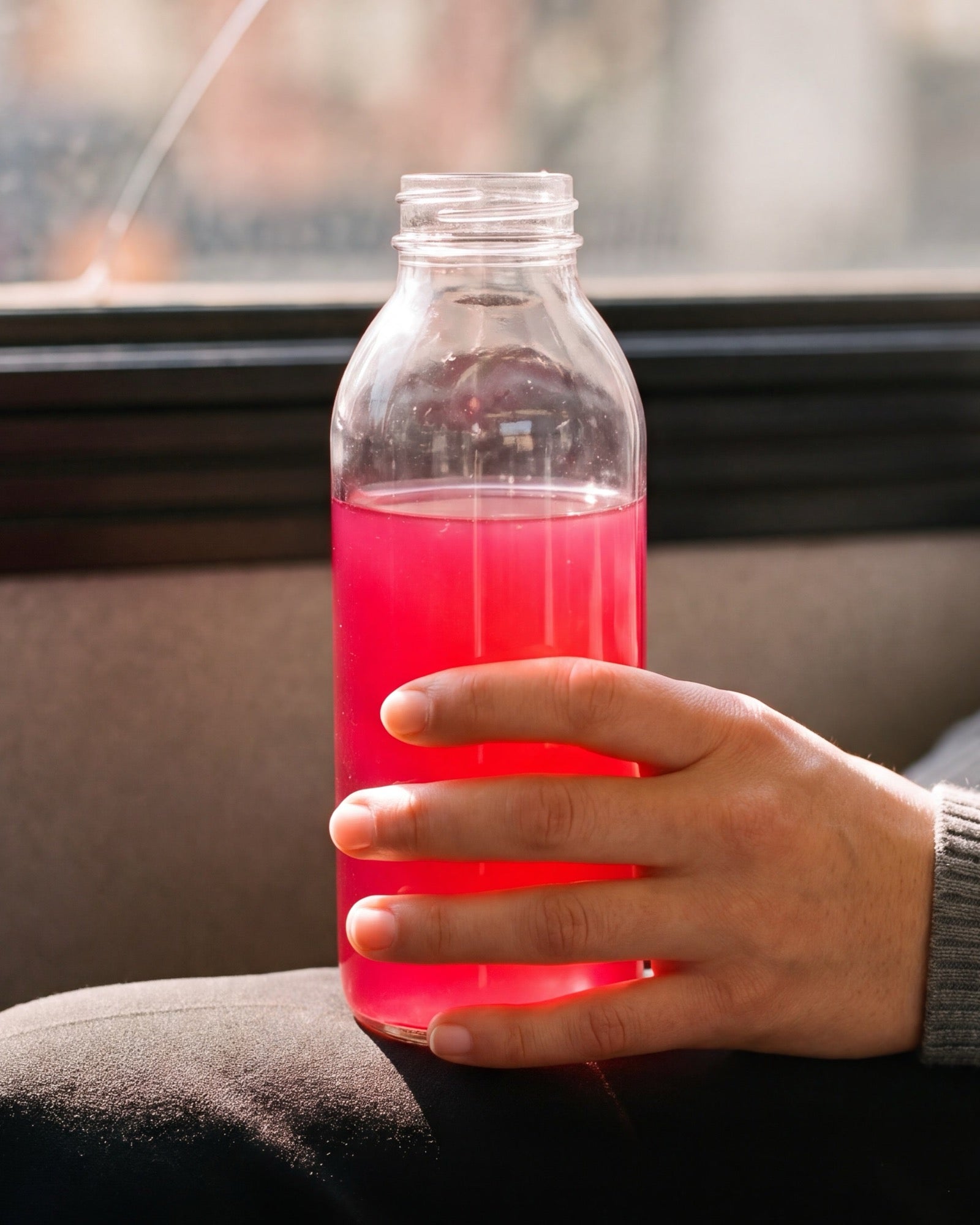 A hand resting around a clear glass bottle of bright pink Desert Drip, warm window light behind