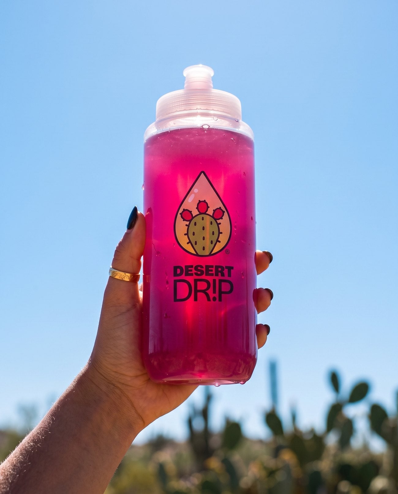 A hand holding a bottle of bright pink Desert Drip against a clear desert sky