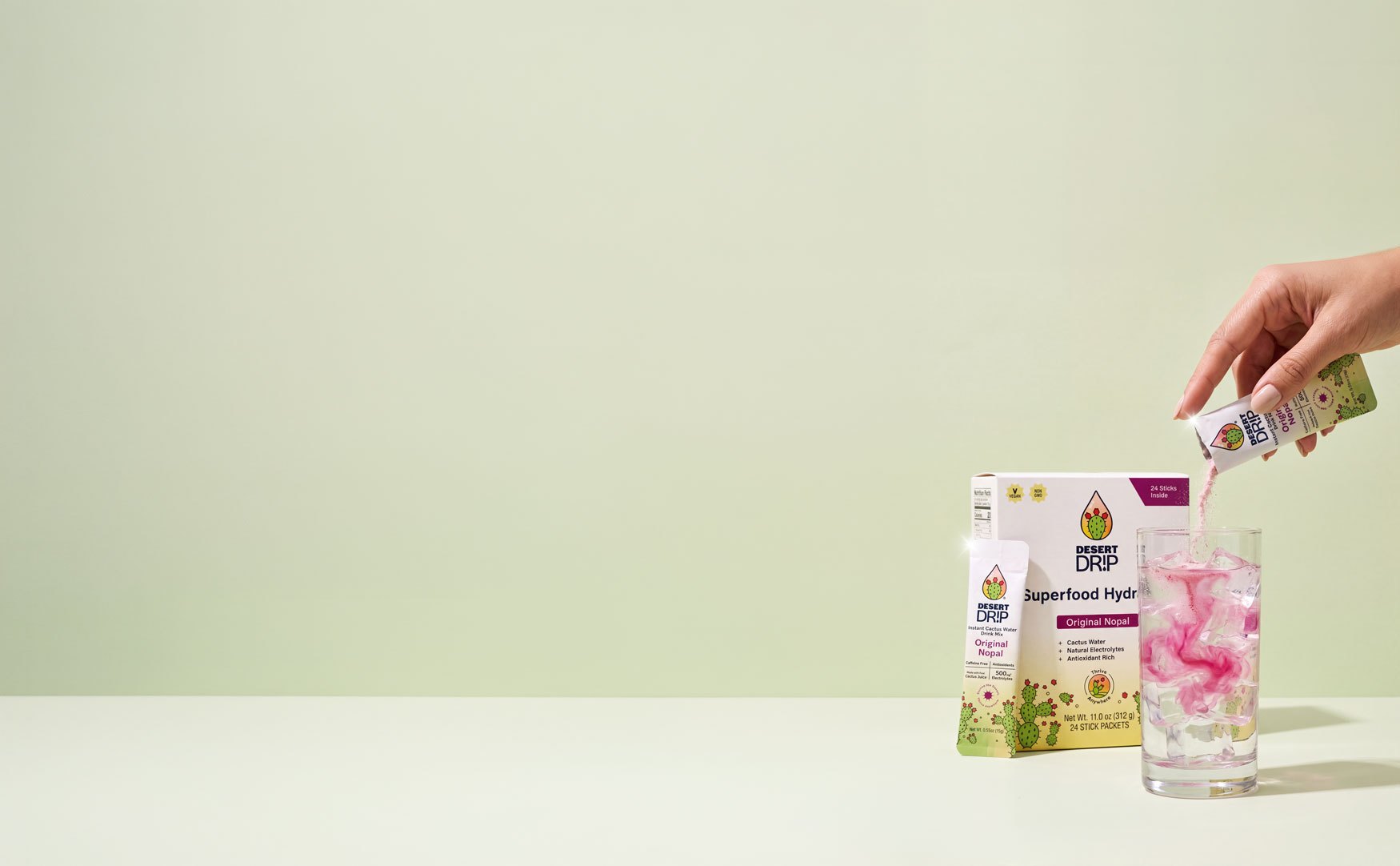 A hand tearing a Desert Drip stick over an iced glass beside the 24-stick box, on a soft green background
