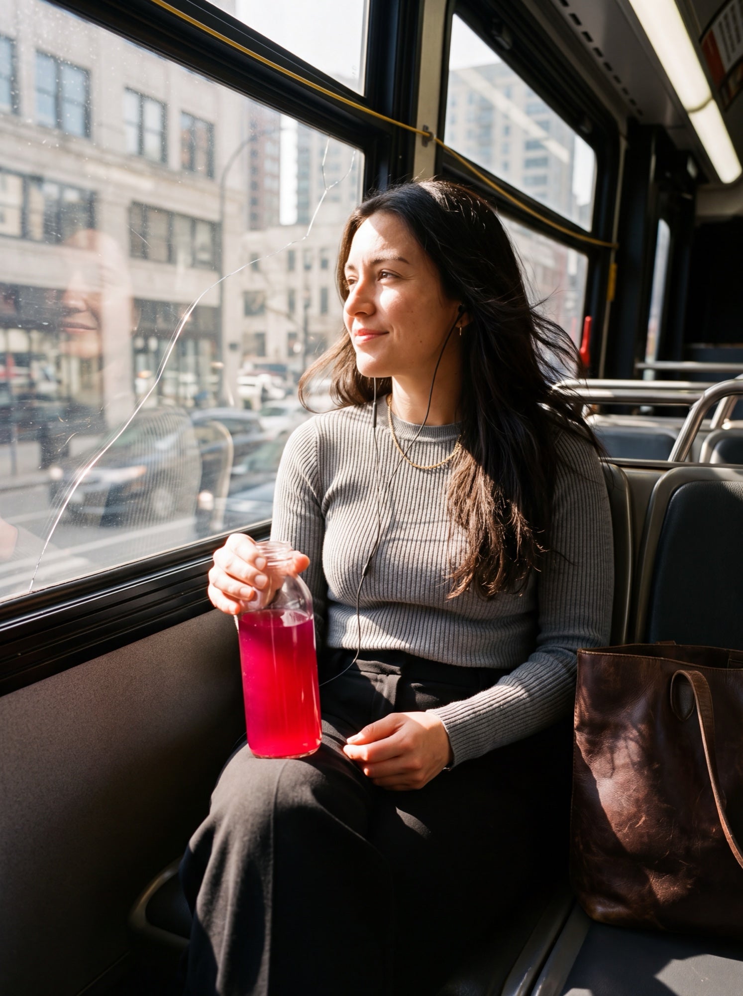 A woman by the window on a city bus, earbuds in, holding a bottle of bright pink Desert Drip