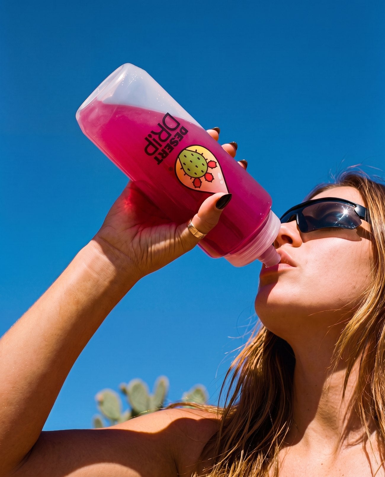A woman outside drinking a bright pink Desert Drip from a shaker against a clear blue sky